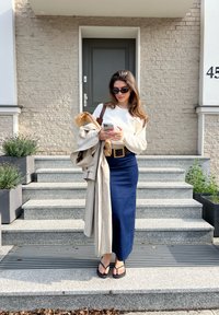 Ženska v kremni dukserici in modri kavbojskih krilih drži pametni telefon. Nosila je japonke in imela na sebi bež plašč. Stopnice so iz sivo kamenja.