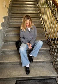 Grå långärmad skjorta, ljusblå vida jeans, svarta loafers och en svart handväska. Sitter på stentrappor med en strukturerad yta.