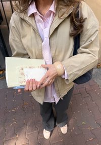 Beige zip-up jacket over a light pink striped shirt, grey trousers, and light shoes. Holding notebooks and a pen with a large translucent bracelet.