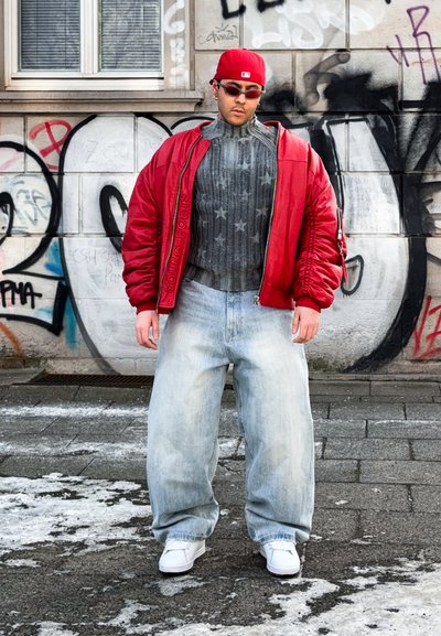Joven con una gorra roja, chaqueta bomber roja, suéter gris con estampado de estrellas, jeans sueltos claros y zapatillas blancas de pie en una calle cubierta de grafitis.