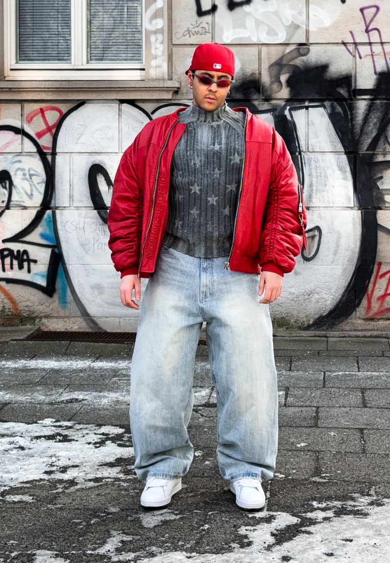 Jeune homme portant une casquette rouge, une veste bomber rouge, un pull gris à motifs d'étoiles, un jean ample clair et des baskets blanches, debout dans une rue couverte de graffitis.