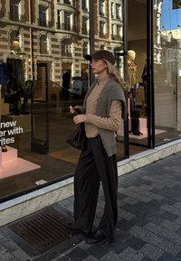 Modell iført en beige kabelstrikket genser, grå ermeløs vest og mørke vide bukser, holder en brun håndveske utenfor en butikk.