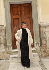 Robe longue noire avec texture florale, associée à un trench-coat crème. Accessoire : un sac beige. L'arrière-plan présente des portes en bois et un mur en pierre.