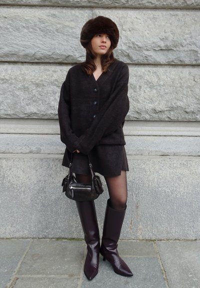 Mujer con cárdigan oscuro de gran tamaño, falda a juego, sombrero de piel marrón, bolso negro y botas altas de color púrpura oscuro, de pie contra una pared de piedra.