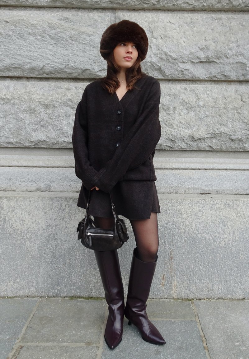 Femme portant un cardigan sombre surdimensionné, une jupe assortie, un chapeau en fourrure marron, un sac à main noir et des bottes hautes violettes foncées, debout contre un mur en pierre.