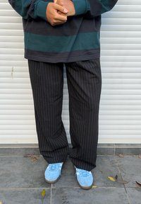 Personne portant un pantalon noir à fines rayures, des baskets bleues, et une chemise à manches longues rayée vert foncé et noire, debout sur un sol carrelé avec des feuilles.