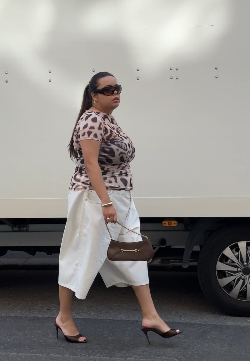 Femme en haut imprimé animal, jupe blanche et talons hauts marchant à côté d'un grand véhicule blanc tenant un sac à main marron avec une chaîne dorée.