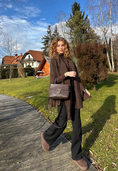 Cárdigan marrón de mangas largas, pantalones negros y zapatos marrones, llevando un bolso oscuro. El fondo presenta árboles y una cabaña de madera.