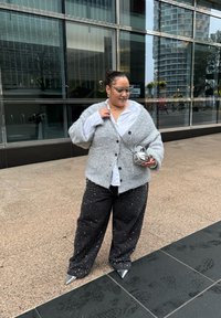 Grijze gebreide cardigan met grote knopen over een wit overhemd, gecombineerd met losse zwarte broek met zilveren studs en puntige metalen hakken.