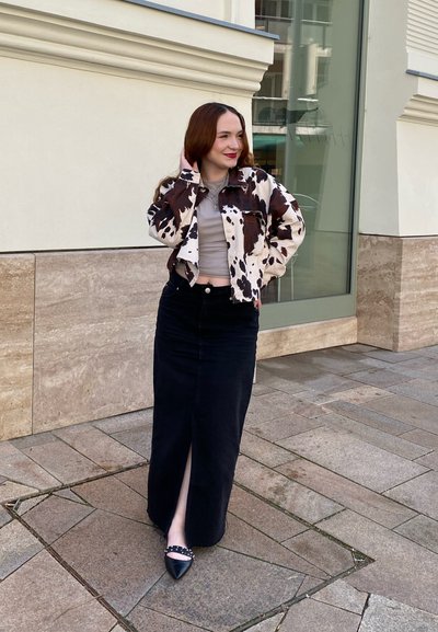 Chaqueta de estampado de vaca marrón y blanca, top corto gris, falda de mezclilla negra con una abertura frontal, zapatos planos negros con acentos de tachuelas.