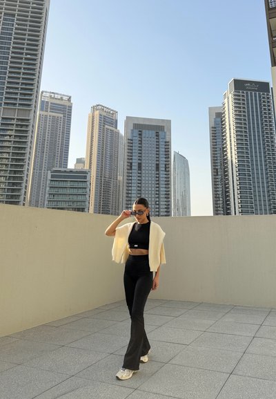 Mujer con top corto negro, pantalones negros, zapatillas blancas y suéter color crema sobre los hombros ajustándose las gafas de sol en una azotea con el horizonte de la ciudad de fondo.