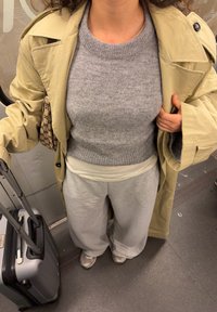 Trenci bej, pulover tricotat gri, pantaloni largi gri deschis și adidași argintii. Geantă neagră și maro cu model. Valiză cu roți în apropiere.