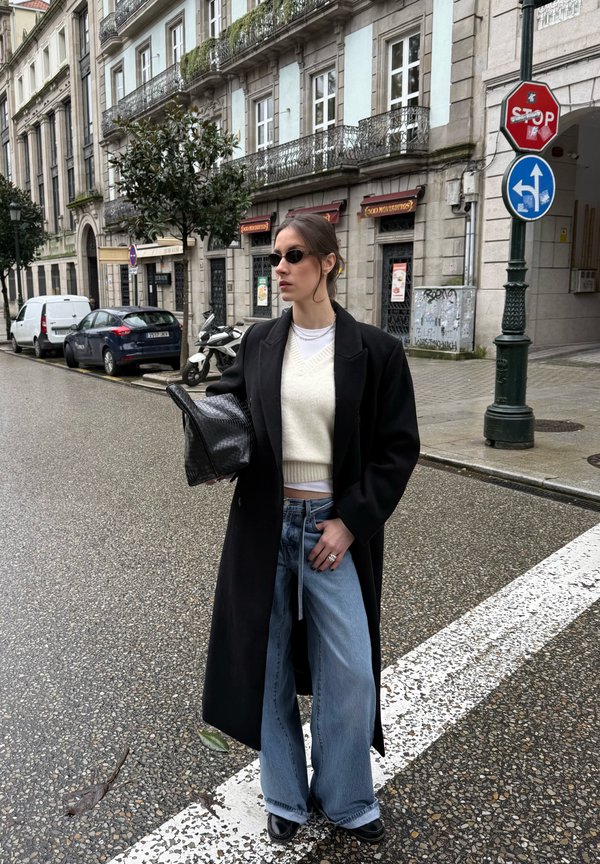 Femme portant un manteau noir, un pull blanc, un jean bleu et des lunettes de soleil, debout à un coin de rue, tenant un sac noir texturé près de bâtiments historiques.