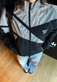 Personne portant une veste Reebok noire et grise, un jean bleu clair et des chaussures noires et blanches, debout sur un sol en bois.