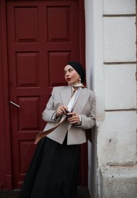 Ženska v sivi blazer, črni ruti in dolgem črnem krilu se opira ob belo steno, drži fotoaparat in stoji ob maroon obloženih vratih.