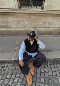 Homme portant un gilet noir, une chemise blanche, un pantalon noir, des bottes marron et une casquette noire avec des cache-oreilles gris, assis sur le bord d'un trottoir en pavés.