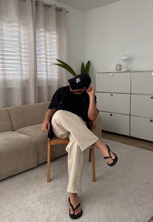 Silla de madera con asiento trenzado, sofá beige, cortinas gris claro, gorra negra, camisa negra, pantalones crema y chanclas negras sobre el suelo alfombrado.