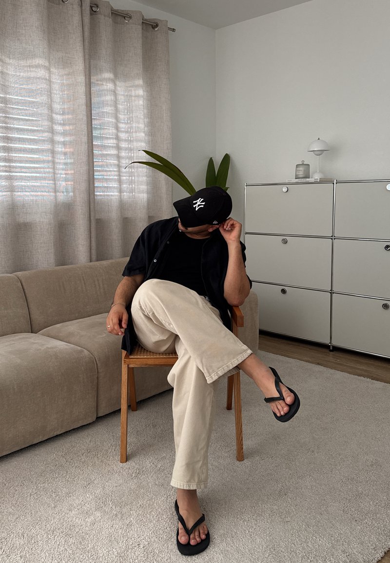 Holzstuhl mit geflochtenem Sitz, beiger Sofa, hellgraue Vorhänge, schwarze Kappe, schwarzes Hemd, cremefarbene Hose und schwarze Flip-Flops auf dem Teppichboden.