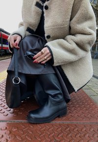 Stivali neri alti fino al ginocchio con una texture in pelle liscia, indossati con un cappotto beige peluche e una gonna in pelle scura, posizionati su una piattaforma con motivo.