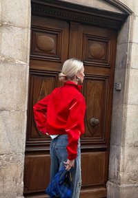 Femme blonde en veste rouge et jean tenant un sac bleu, debout devant une grande porte en bois ornée encadrée par des murs en pierre.