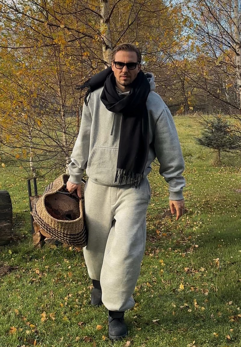 Felpa grigia con cappuccio e pantaloni della tuta, sciarpa nera. Tiene un cesto intrecciato di forma rotonda. Foliage autunnale sullo sfondo. Area erbosa verde.