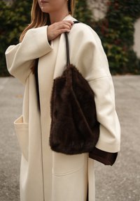 Brązowa torebka z futerkiem syntetycznym o miękkiej fakturze, trzymana za czarny pasek. W tle zielone liście i neutralna betonowa powierzchnia.