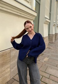 Tmavomodrý pletený kardigán s výstrihom do V a zapínaním na gombíky, s dlhými voľnými rukávmi. Dopĺňajú ho tmavosivé džínsy s širokými nohavicami.