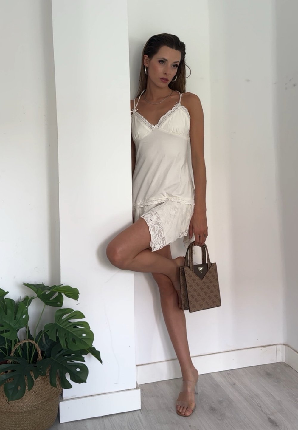 Abito bianco a canotta con pizzo, abbinato a una borsa marrone texturizzata. Foglie dimensionali in un cesto intrecciato nelle vicinanze. Sfondo chiaro.