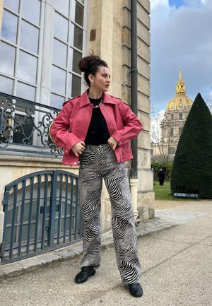 Chaqueta corta rosa, top negro, pantalones de pierna ancha de estampado de cebra y zapatos negros. De pie cerca de un edificio con grandes ventanas y una cúpula en el fondo.