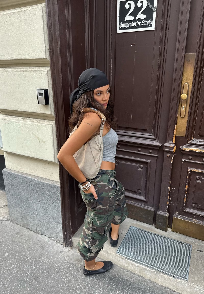 Donna che indossa un top corto grigio e pantaloni verdi con camouflage, mentre tiene una borsa beige chiaro. Sciarpa nera, scarpe nere basse. Porta di legno sullo sfondo.