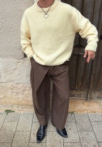 Persoană îmbrăcată într-un pulover crem tricotat, pantaloni largi maro cu dungi fine, cizme negre și un colier cu pandantiv inima argintie, stând pe o pavează de piatră.