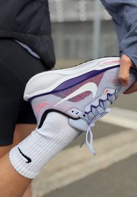 Szare buty sportowe z wielokolorową siateczkową cholewką, białą podeszwą, fioletowymi akcentami i czerwonymi detalami. Noszone z białą skarpetą ze wzorem prążków i czarnym logo.