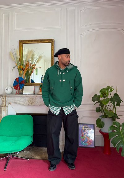 Hombre con sudadera verde con capucha, boina negra y pantalones negros sueltos está de pie en una habitación con una silla verde, plantas, obras de arte enmarcadas y una chimenea ornamentada.