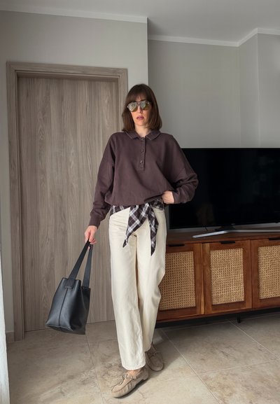 Mujer con gafas de sol, camisa de manga larga de color marrón oscuro, camisa de cuadros atada a la cintura, pantalones crema, sosteniendo un bolso negro tipo tote en el interior cerca de una puerta de madera y una consola de televisión.