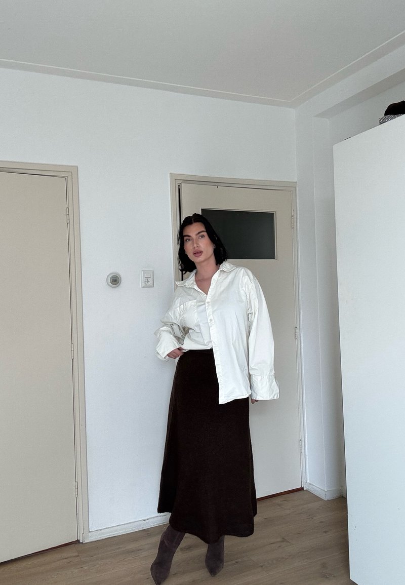 Femme aux cheveux noirs portant une chemise blanche oversize, une longue jupe marron foncé et des bottes assorties, debout à l'intérieur contre des murs blancs et des portes fermées.