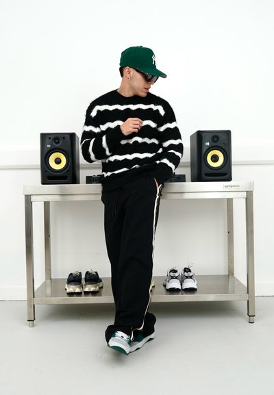 Suéter difuso a rayas blancas y negras, pantalones negros con rayas blancas, gorra verde, zapatillas negras con detalles amarillos, zapatillas exhibidas.