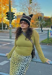 Lange mouw olijfgroene top, geel patroonrok met zwarte vlekken en een gebreide muts met puntige oren. Accessoires omvatten een schoudertas.