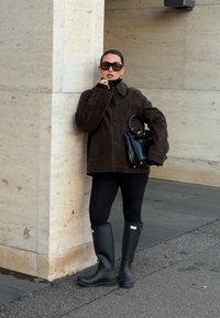 Brun semsket jakke med krage, kombinert med svarte leggings og svarte gummistøvler. Holder en liten svart håndveske med gullbeslag.