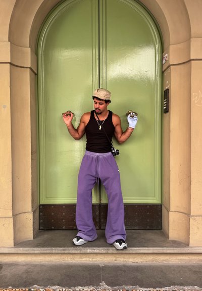 Persona vestida con una camiseta sin mangas negra y pantalones de chándal morados, sosteniendo pomos de puertas, de pie frente a una puerta doble verde con un acabado texturizado.