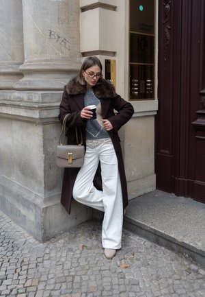 Mujer con un abrigo largo oscuro con cuello de piel, pantalones blancos y gafas, de pie en una calle empedrada sosteniendo una taza de café y un bolso beige.