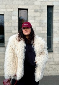 Casaco de pelúcia falso de cor creme com uma textura desgrenhada, usado por cima de um hoodie preto. Uma touca cor bordeaux é usada em cima, e uma pequena bolsa vermelha é segurada.