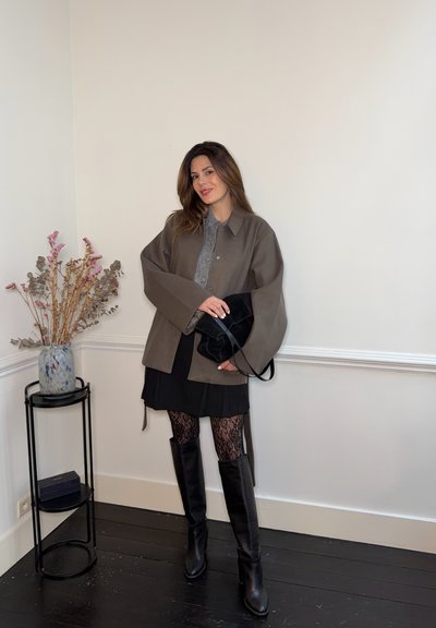 Mujer de pie en el interior, vestida con una chaqueta marrón, falda negra, medias de encaje y botas negras hasta la rodilla, sosteniendo un bolso negro junto a un soporte para plantas.