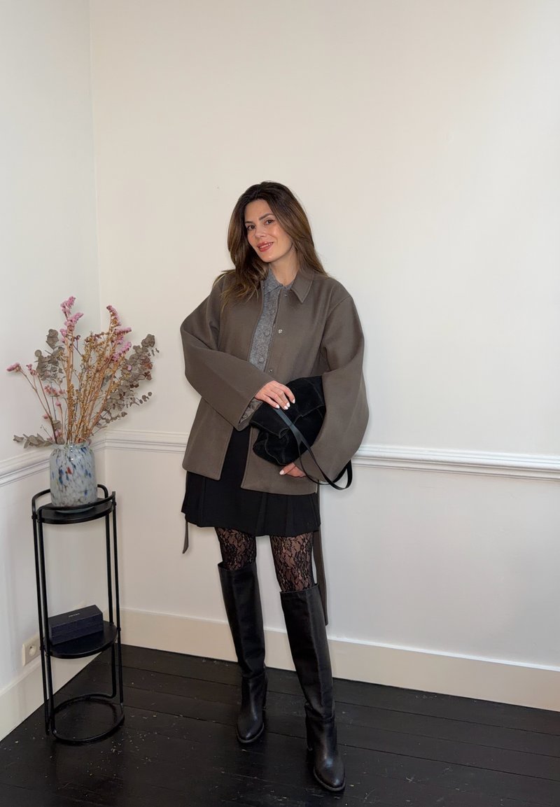 Donna in piedi all'interno, indossa una giacca marrone, gonna nera, calze di pizzo e stivali neri al ginocchio, tiene una borsa nera accanto a un supporto per piante.