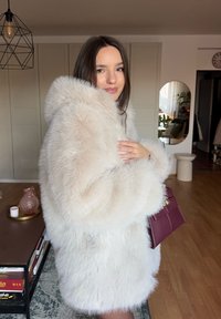 Femme en manteau fausse fourrure crème tenant un sac à main bordeaux dans un salon moderne avec des sols en bois et des objets décoratifs.