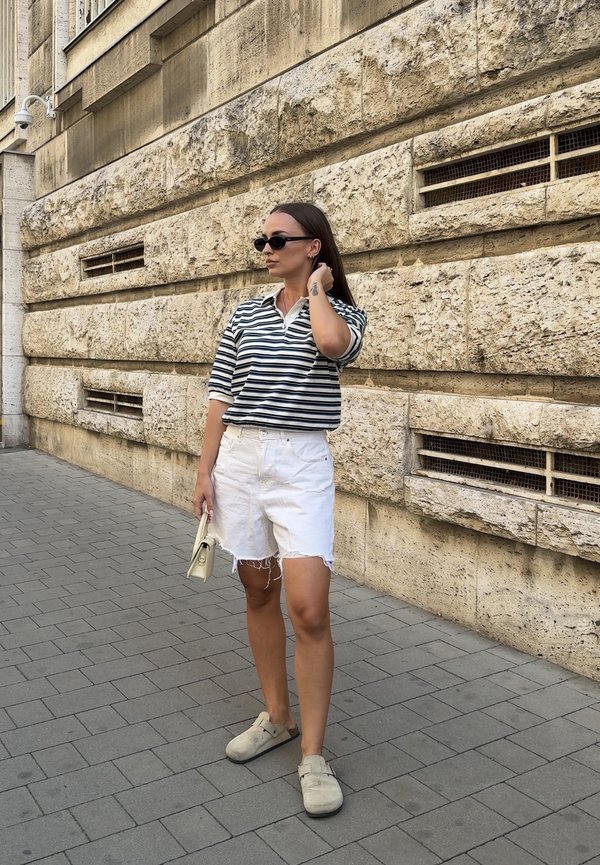 Frau trägt ein schwarz-weiß gestreiftes Shirt, weiße Shorts und Sonnenbrille und steht auf einem gepflasterten Gehsteig neben einer Steinmauer.