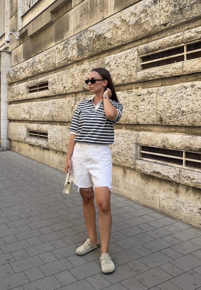Femme portant un t-shirt rayé noir et blanc, un short blanc et des lunettes de soleil, se tenant sur un trottoir pavé à côté d'un mur en pierre.
