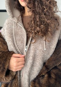 Nepbont poncho in lichtbeige met een bruine, ruige buitenkant. Voorzien van een capuchon, ritssluiting en trekkoorden. Zachte, getextureerde stof over het geheel.