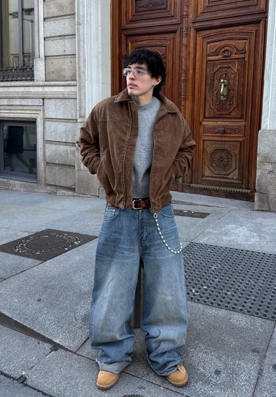 Joven con cabello rizado y oscuro, lleva gafas, chaqueta marrón, suéter gris, vaqueros holgados con una cadena y botas color beige, de pie en la acera de la ciudad.