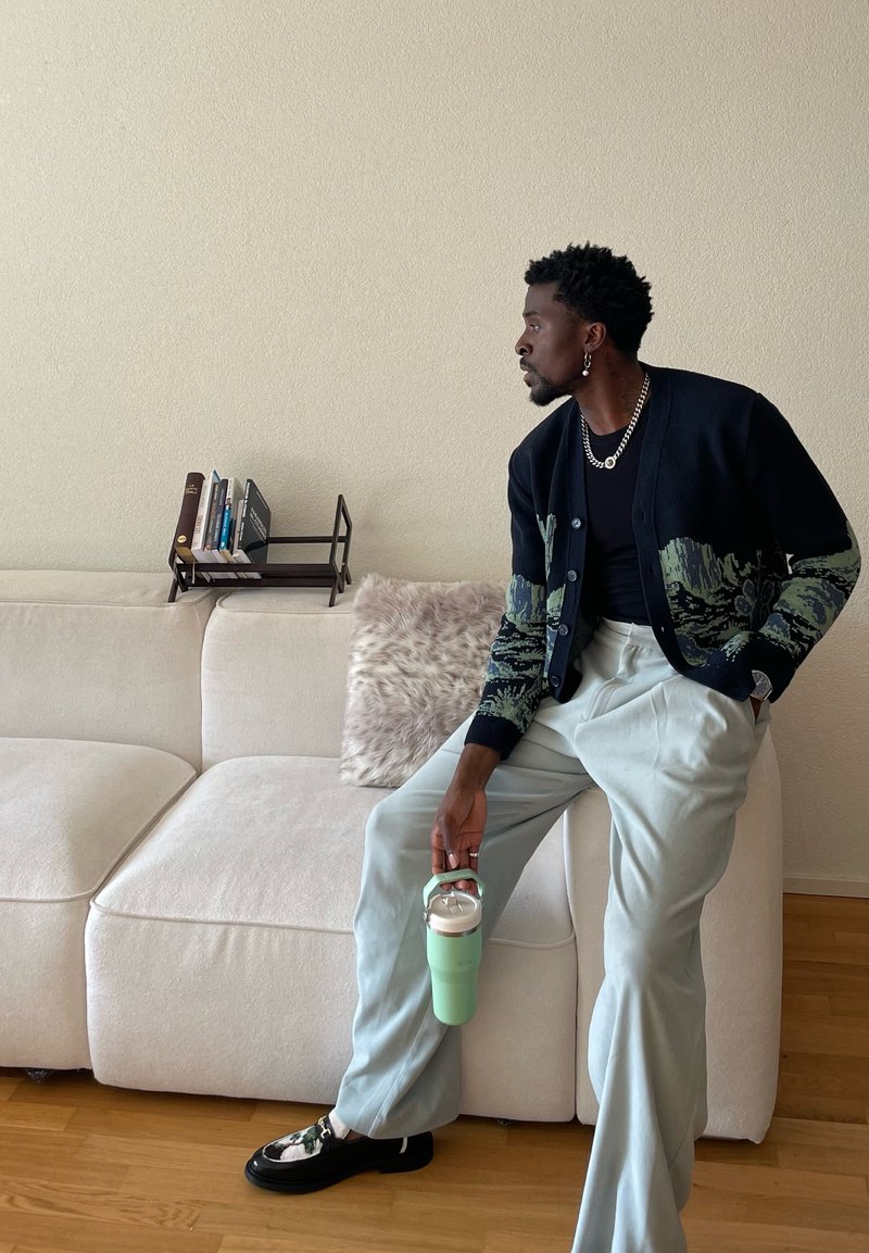 Homme en cardigan à motifs et pantalons larges tenant une bouteille verte, assis sur un canapé beige avec des livres et un coussin en fourrure derrière lui.