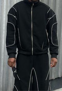 Conjunto atlético negro con chaqueta con cremallera y pantalones, con un patrón de costuras blancas. La tela tiene una textura suave y se muestra sobre un fondo gris.